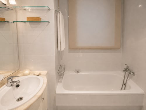 Baño limpio del hotel Holibai en Baiona con paredes de azulejos blancos, lavamanos, bañera y estantes de vidrio