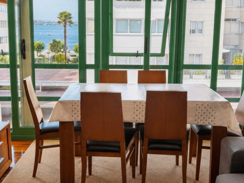 Comedor de Holibai en Baiona con mesa de madera, sillas marrones y vistas al mar a través de ventanas con marco verde