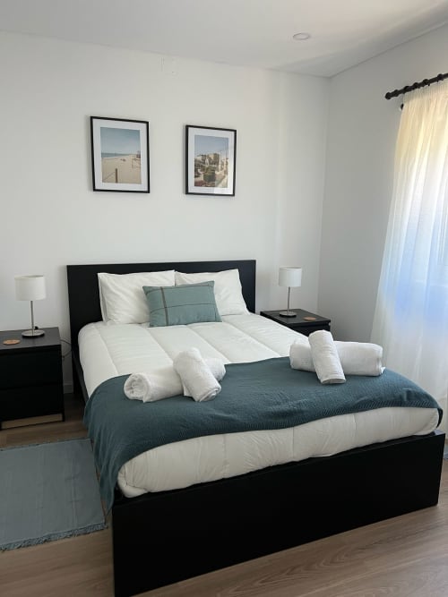 Quarto moderno da villa Casa do Sol Norte com cama preta, roupa de cama branca, toalhas enroladas e arte costeira emoldurada acima