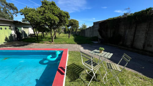Piscina clara com água turquesa e borda vermelha, espreguiçadeiras, gramado verde, árvores e paredes de concreto na Casa do Sol