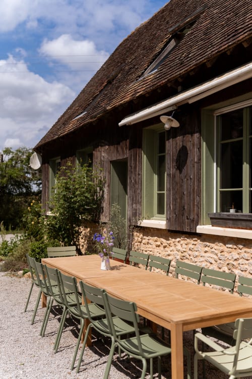 Terrasse de restauration en plein air aux Maisons Noisette avec table en bois, chaises vertes, cottage en pierre rustique et ciel bleu