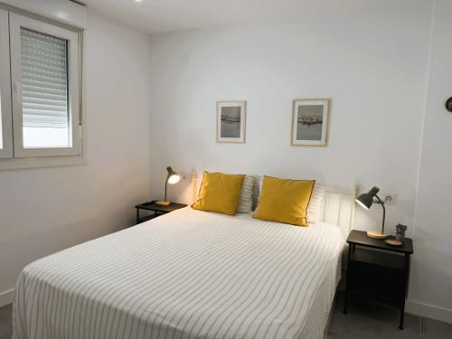 Dormitorio moderno con ropa de cama a rayas blancas, cojines amarillos, mesillas negras y cuadros enmarcados en Fuengirola de lujo