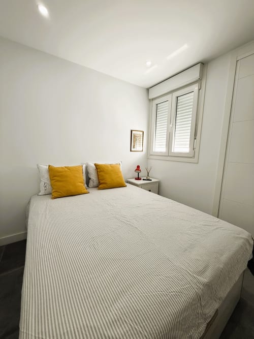 Luminoso dormitorio moderno en lujoso apartamento de Fuengirola con cama blanca, almohadas amarillas, ventana con persianas y decoración minimalista