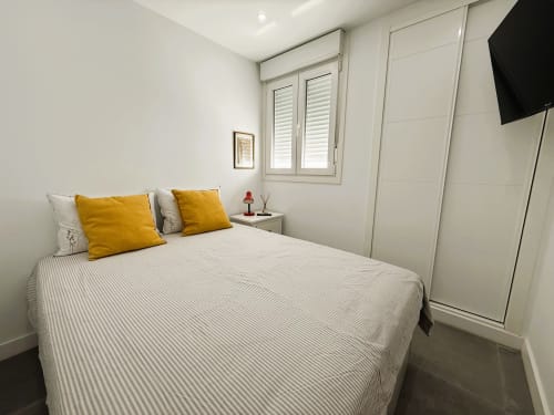 Moderno dormitorio con cama de matrimonio, almohadas amarillas, ventana con persianas blancas, TV mural y armario empotrado en Fuengirola