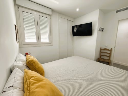 Dormitorio moderno en lujoso apartamento frente al mar, Fuengirola. Cama queen con almohadas amarillas, TV montada en pared, persianas blancas