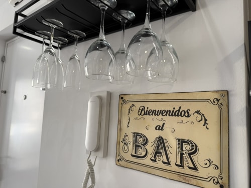 Copas de vino colgadas de una rejilla negra sobre cartel vintage 'Bienvenidos al BAR' en lujoso apartamento en la playa de Fuengirola