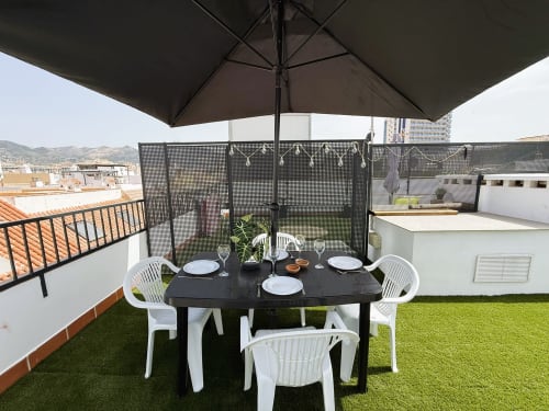 Terraza en azotea con comedor en apartamento de Fuengirola: mesa negra, sillas blancas, sombrillas, césped artificial y vistas a la ciudad