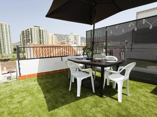 Terraza de lujo en azotea con zona de comedor, césped artificial, sombrilla negra, sillas blancas y vistas a la ciudad de Fuengirola