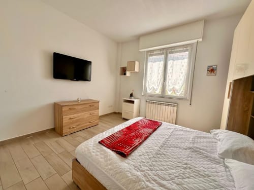 Camera luminosa al Welcome Holiday di Loano con letto bianco, cassettiera in legno, TV e finestra con vista sul giardino