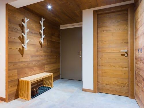 Entrée contemporaine des Tanières avec murs en bois, porte-manteaux blancs, banc d'accueil et porte d'entrée grise