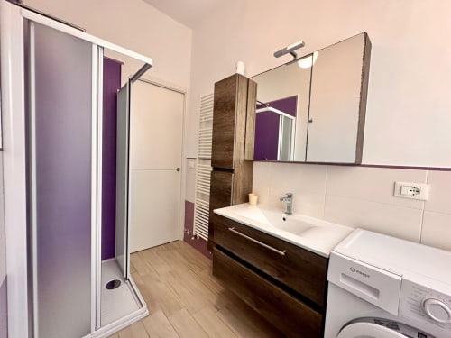 Bagno moderno nell'appartamento di Loano con vanity in legno scuro, lavabo bianco, specchio, doccia, lavatrice e pavimento in legno
