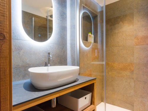 Salle de bain moderne avec miroir ovale lumineux, vasque blanche, meuble en bois et douche à l'italienne avec paroi vitrée