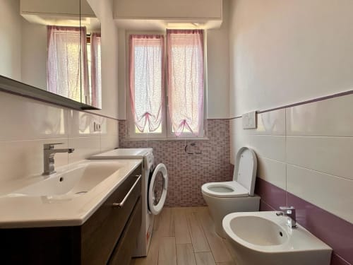Bagno moderno nell'appartamento di Loano con doppio lavabo, wc, lavatrice, tende rosa e pavimento effetto legno