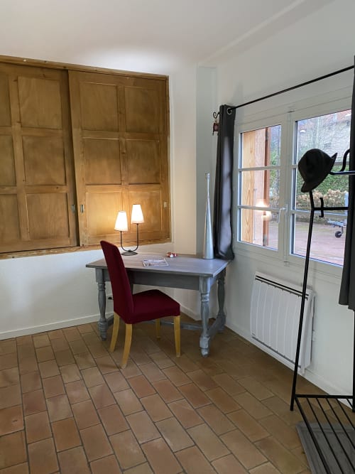 Espace de travail à domicile au gîte Les Vaches avec bureau en bois, chaise bordeaux, lampe et vue sur le jardin par les portes-fenêtres
