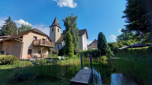 Le gîte LA JUMENT VERTE à Trévignin avec son église, sa jetée en bois, son étang et ses jardins luxuriants sous un ciel bleu