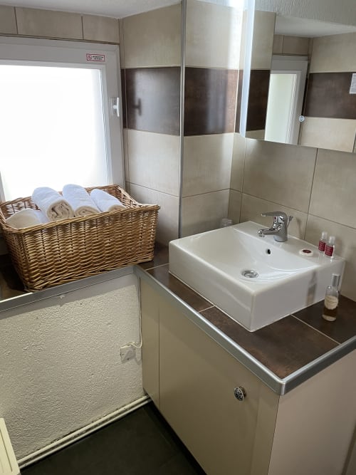 Salle de bain moderne du gîte Les Vaches avec lavabo blanc rectangulaire, carrelage marron et beige, panier à serviettes en osier