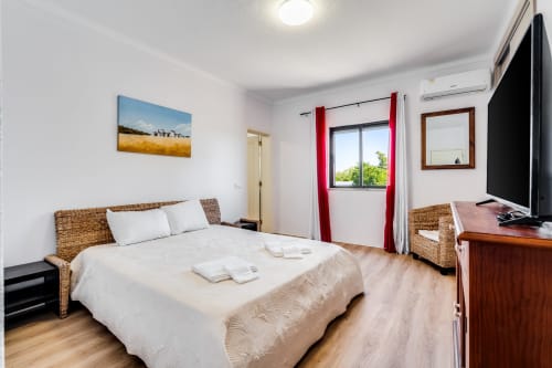 Bright bedroom with wicker bed frame, white linens, flat-screen TV, and red curtains overlooking the garden at Quinta do