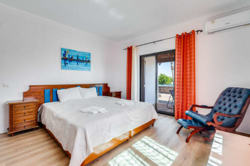 Bright bedroom with wooden bed, white linens, blue accents, orange curtains, and sea view at Quinta do Águas villa in Lagoa,
