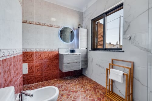 Modern bathroom with red and white tile, round mirror, sink vanity, wooden towel rack, and window overlooking Lagoa, Portugal