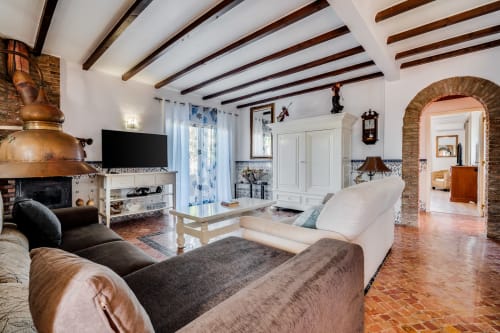 Spacious villa living room with exposed wooden beams, fireplace, TV, terracotta floors, and archway opening to pool area at