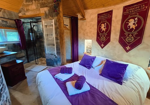 Luxuriöses Schlafzimmer im La Synchronicité B&B in Saint Sauveur mit violetten Samtkissen, mittelalterlichen Bannern und Holzbalken