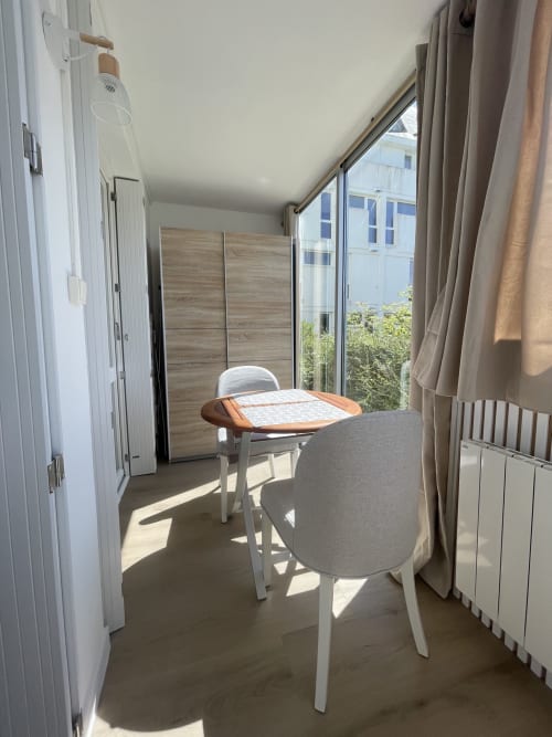 Studio lumineux avec table ronde, deux chaises, armoire en bois, radiateur et vue sur la mer à Guimiliau, Finistère