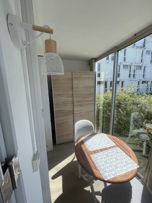 Balcon lumineux d'un petit appartement avec table à manger en bois, chaise blanche, lampe suspendue, écran de confidentialité, plantes en pots et vue sur la ville