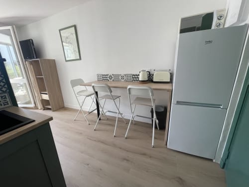 Kitchenette studio avec réfrigérateur blanc Beko, coin repas avec chaises et équipements modernes à Roscoff