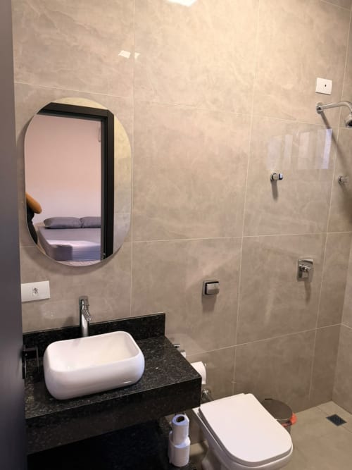 Banheiro moderno com cuba branca, bancada escura, espelho oval e área de duche na pousada Casa Coqueiros em Maringá,
