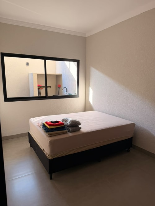 Quarto da pousada Casa Coqueiros em Maringá com cama branca, roupas dobradas, espelho e decoração minimalista