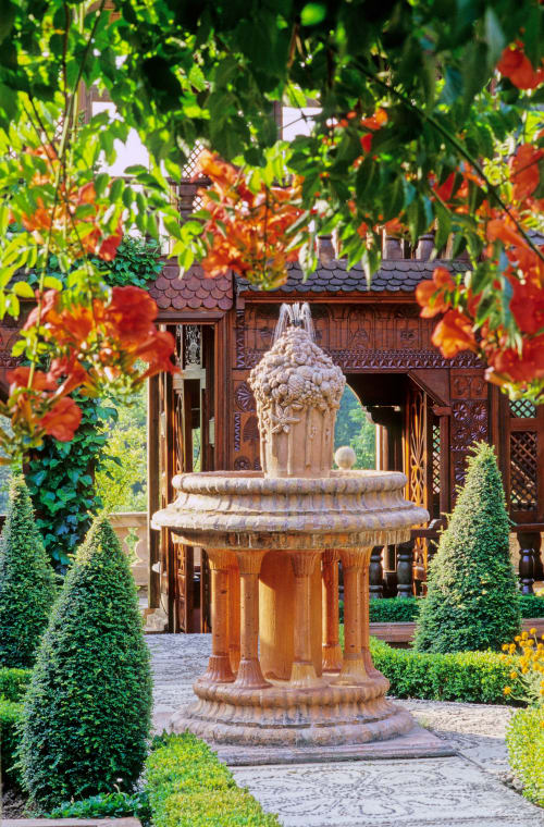 Fontaine ornementale en pierre dans le jardin paysager avec fleurs rouges, topiaires et pavillon en bois aux Micro Palaces B&B, Vaulx, France.