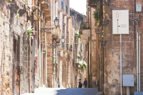 Stretto vicolo storico a Taranto con edifici in terracotta e edera, lampioni e una coppia che passeggia - Zappy