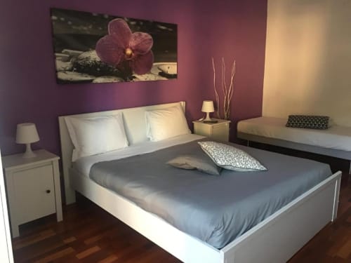 Moderna camera tripla a Domum Vaticani Roma con letto queen, pareti viola, orchidee, comodini bianchi e pavimento in legno