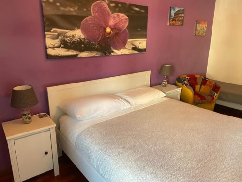 Camera luminosa della Domum Vaticani Roma B&B con letto bianco, muri viola, opera d'arte con orchidea rosa e lampada comodino