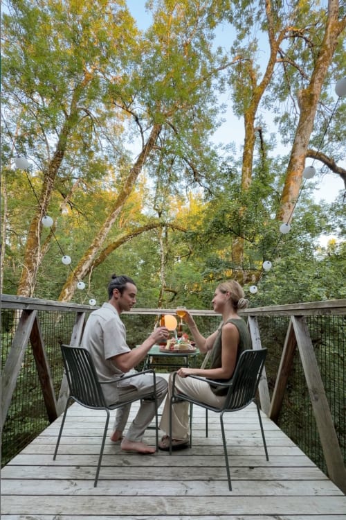 Couple trinquant sur une terrasse en bois sous les grands arbres de l'hôtel-boutique Micro Palaces à Vaulx, France