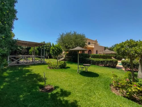 Jardin de villa toscane avec pelouse verdoyante, pergola, table parasol et bâtiment en terre cuite à Villa Castellum