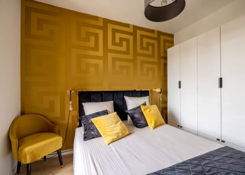 Chambre moderne avec mur d'accent géométrique doré, lit queen avec oreillers jaunes, fauteuil doré, armoire blanche