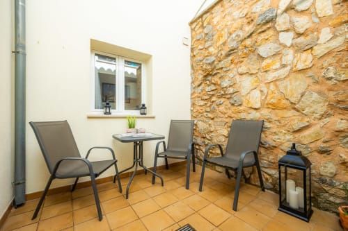 Rústica zona de estar en terraza de apartamento Artà con baldosas de barro, sillas metálicas, mesita y encantador muro de piedra