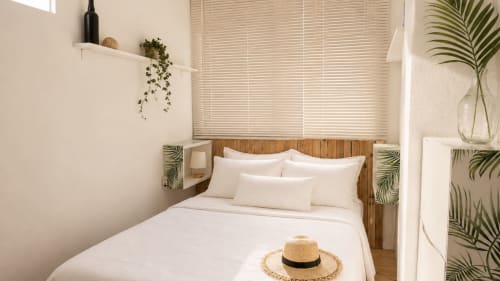 Dormitorio luminoso en Holidays Costa Brava con cabecero de madera, ropa de cama blanca, plantas tropicales y sombrero de paja sobre la cama