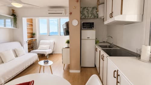 Moderno apartamento estudio en Holidays Costa Brava con cocina integrada, sofá blanco, comedor y vistas panorámicas de la ciudad