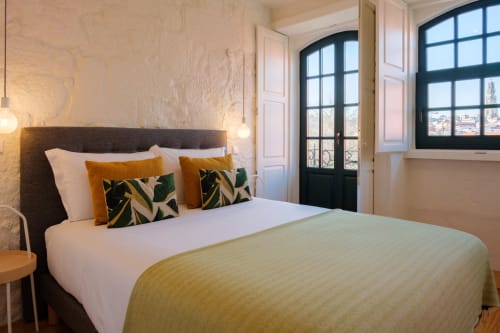 Quarto moderno no AgapeStay Porto com cabeceira escura, roupa de cama em tons de azeite, almofadas estampadas e vista para a cidade