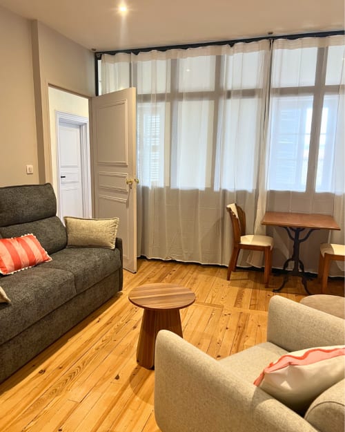 Séjour lumineux à Kipling House avec parquet, canapé gris, fauteuil, table d'appoint en bois et grandes fenêtres voilées