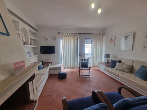 Sala de estar moderna e luminosa no apartamento Solazur em Ericeira, com piso em terracota, móveis confortáveis, TV e portas envidraçadas