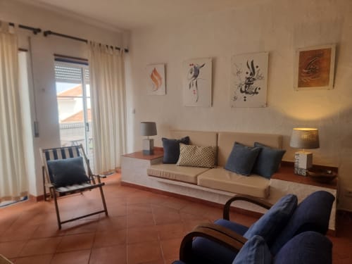 Ampla sala de estar do apartamento Solazur em Ericeira com vistas da cidade, sofá bege, poltronas azuis e decoração moderna.