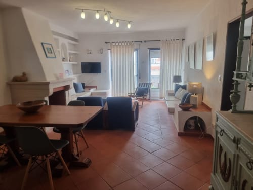 Espaçosa sala de estar do apartamento em Ericeira com mesa de jantar, móveis, cozinha, lareira e vista para a cidade pelas portas do varanda