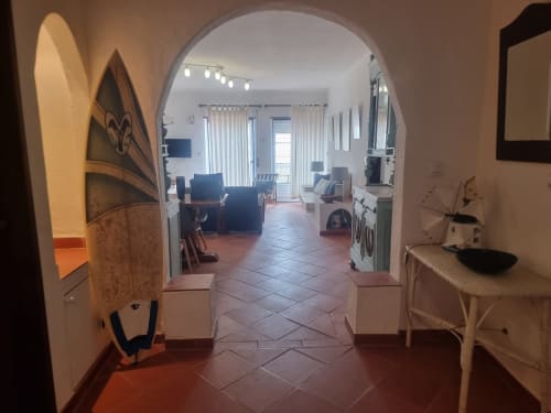 Corredor do apartamento Solazur em Ericeira, levando à sala de estar clara com porta em arco, decoração com prancha de surfe e mobiliado