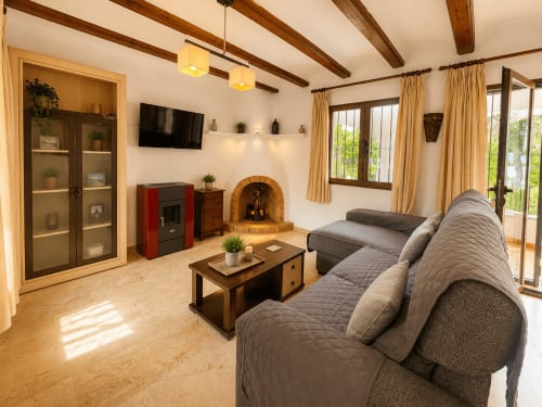 Amplio salón de villa en Moraira con sofá seccional gris, chimenea, TV, vigas de madera y vistas al jardín