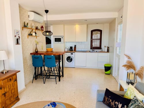 Moderna zona de cocina y salón en apartamento de Nerja con taburetes azules, muebles blancos y planta abierta