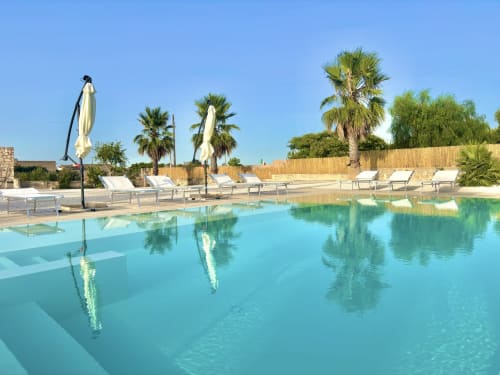 Piscine de l'hôtel B&B Helios à Nardò avec eau turquoise, chaises longues, palmiers et parasols sous ciel dégagé