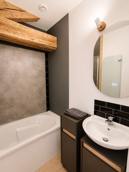 Modern bathroom with wooden beams, round mirror, white sink, and shower tub at Domaine de Beau Site cottage in